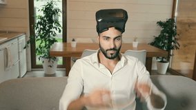 Handsome bearded arab man wears white shirt uses VR helmet with futuristic visual interface, slide gestures on hologram control panel sitting in the kitchen