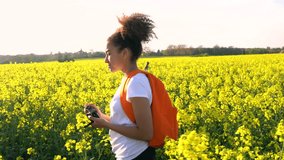 4K video clip of healthy mixed race African American girl teenager female young woman with red backpack and camera taking photograph in field of rape seed yellow flowers