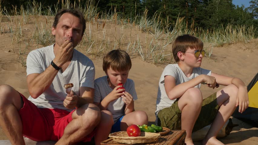 Happy father and two little sons having lunch on a sandy beach, They eating grilled sausages with tomatoes and cucumbers, Closeup, 4k shot