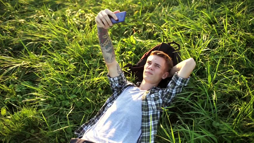 A handsome guy is lying on the grass and talking on video. The student is lying on the grass and talking on the phone. A young man smiles and takes a selfie. Nice guy lying on the grass with his phone