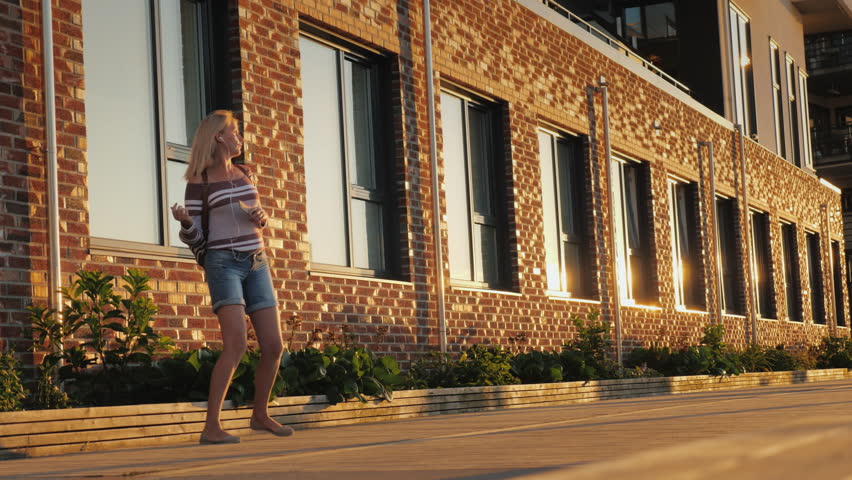 A carefree middle-aged woman listens to music and dances on the street near a beautiful brick building