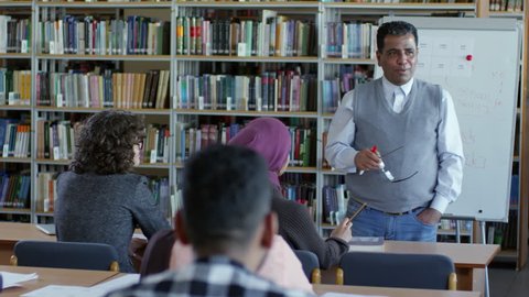 Tilt down of experienced middle eastern male teacher explaining lesson to migrant students in classroom with book racks and flipchart