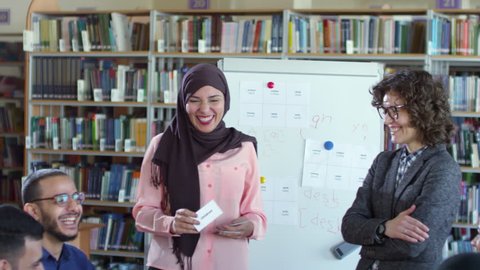 Beautiful muslim woman in hijab standing by flipchart in classroom, smiling and showing word cards to group of migrant students at English lesson with female teacher