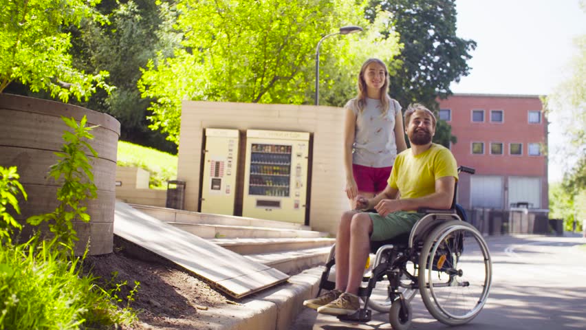 Happy young disable man in a wheelchair in the park with his wife. They showing something by gestures and smiling