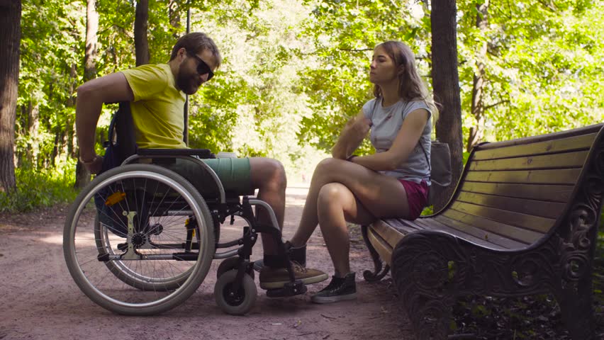 Young disable man in a wheelchair on a walk in the park with his wife. A woman is sitting on a bench. Man smiling