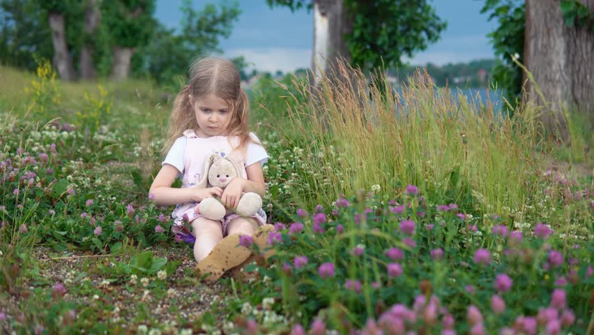A little pretty girl plays with a plush rabbit, she sits in a meadow among a flowering clover.