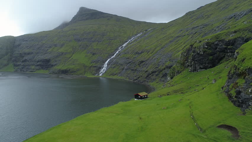 AERIAL: Lonely little house hiding below the steep mountains in the vast green Scandinavian wilderness looking over the beautiful deep blue lake. Flying towards a mountain stream rushing past a house.