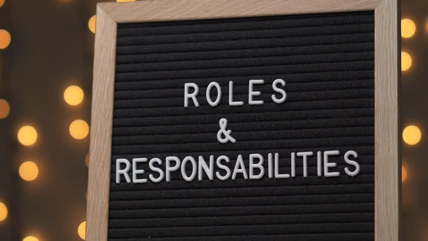 Black letter board with ROLES & RESPONSABILITIES Written on it with white letters. Camera rotating around the sign showing the beautiful bokeh balls in the background. Royalty-Free Stock Footage #1015157443