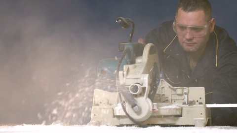 A worker in overalls on a blue background cuts out a 4K wood item on the electric saw. A stream of sawdust from the machine tool when processing a tree. The shavings fly to the camera with smoke.
