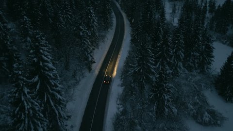 Aerial shot of a car driving next to a snowy forest at dusk.