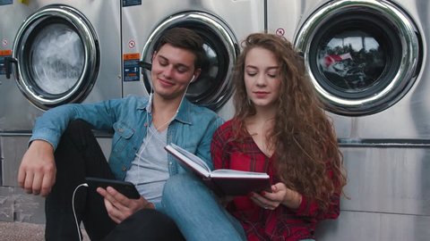 Beautiful couple in a laundry listen to the music on the phone, reading book, watching videos. Handsome young man with stylish hair?ut in jeans shirt. Woman with curly red hair in tartan shirt.