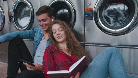 Beautiful couple in a laundry listen to the music on the phone, reading book, watching videos. Handsome young man with stylish haircut in jeans shirt. Woman with curly red hair in tartan shirt.