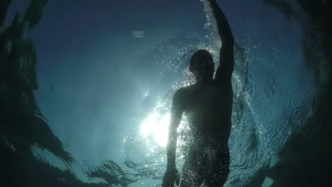 SLOW MOTION CLOSE UP, UNDERWATER, LOW ANGLE VIEW: Muscular guy swimming front crawl freestyle stroke, waterdrops falling and splashing on glassy water surface in amazing blue ocean on sunny summer day