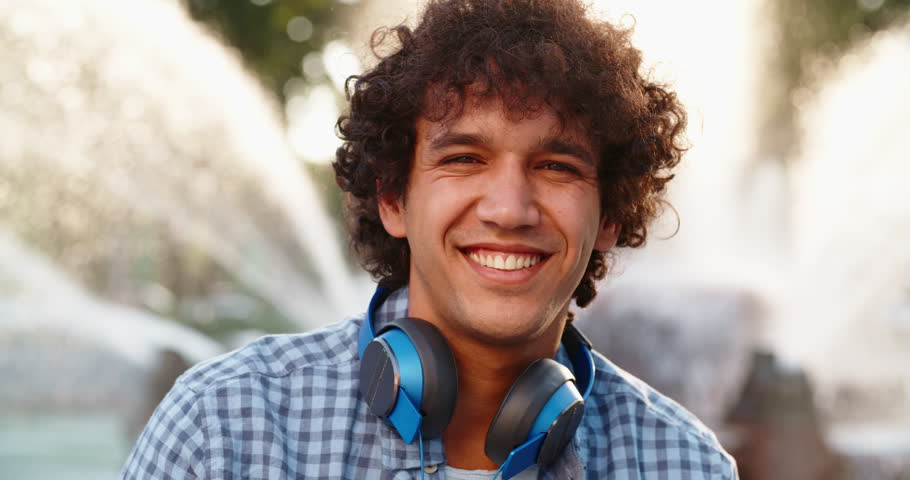 Portrait of young asian guy with curly hair in earphones listening to music and looking at camera 4k