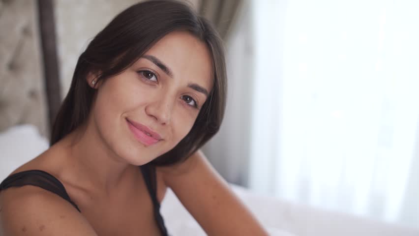 A close-up side view of young brunette's face. She is looking into camera, smiling, and leans her head on the hand, touches her hair and turns the head to the right. | Shutterstock HD Video #1015186963