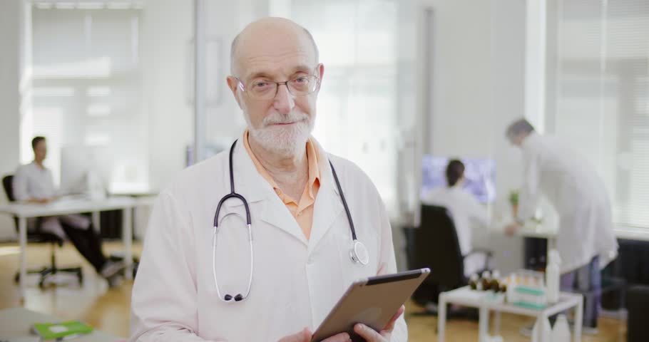 Elderly gray-haired man with glasses, stethoscope and tablet in his hands is in the clinic. Serious male physician therapist looking at the camera