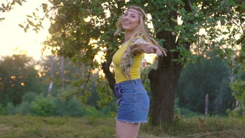 young attractive woman in a yellow T-shirt is spinning at sunset. green apple orchard on background. happy and free hippies