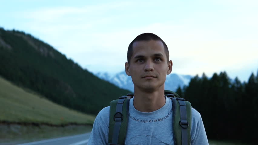 A tourist hitchhiker walks along the asphalt road with a backpack on his shoulders. Ahead is a snowy peak of the mountain.