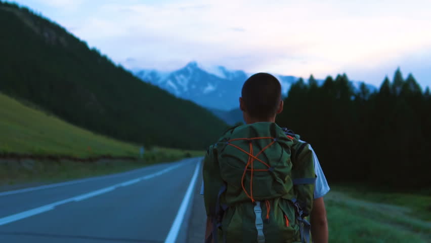 A tourist hitchhiker walks along the asphalt road with a backpack on his shoulders. Ahead is a snowy peak of the mountain.
