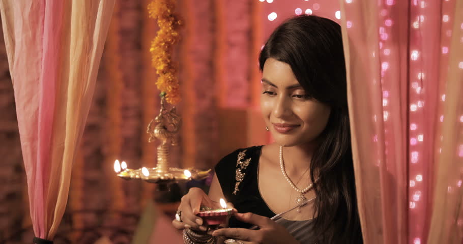 A beautiful woman lighting a lamp (Diya) during Diwali festival. A charming and happy female in a traditional clothing  lighting a hanging metal lamp in a house decorated with lights and flowers Royalty-Free Stock Footage #1015209853