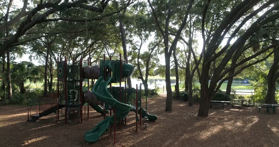 Ground-level view frames a shaded park playground beneath mature trees with slides, climbing equipment, benches, and nearby water, 2018.