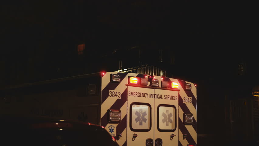 View of rear doors and flashing lights of emergency service ambulance moving away from accident scene at nighttime.