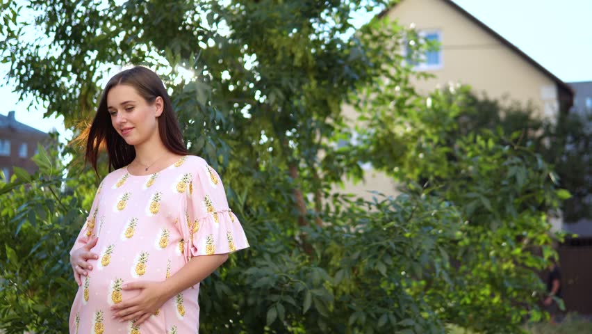 Beautiful pregnant girl in a pink dress stroking her tummy at sunset.
