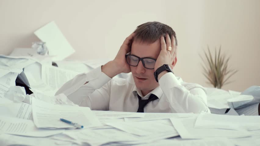 A young tired man in glasses sits in a pile of papers and signs documents. Because of fatigue, a person is upset and depressed. He squeezed his head in his hands. | Shutterstock HD Video #1015280167