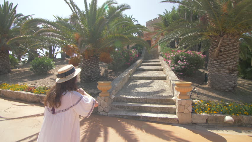 Beautiful woman running up the stairs near luxury villa in Italy. Nice hotel territory. Girl walking and dressed in elegant white dress and hat. Slow motion. Good weather, blue sky. Following camera Royalty-Free Stock Footage #1015295395