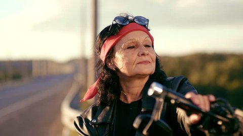 An elderly woman in a red scarf, leather jacket and gloves stands on the background of an empty road. Puts his hands on his chest and shows the sign of bikers. The woman is smiling. Happy life of an