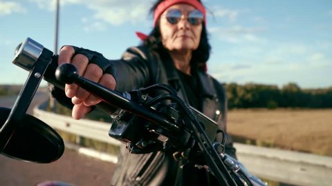 The old biker sits at the wheel of his motorcycle. On a steep motorcycle sits an elderly woman in round glasses, she has a red bandana and a leather jacket, and gloves. An old biker with a nature
