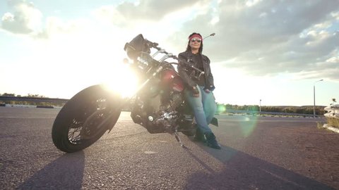 Biker old woman in leather jacket and gloves sitting on his cool motorcycle. The woman has round glasses and a red bandana. In the background is an empty road and a beautiful evening. Biker shows sign