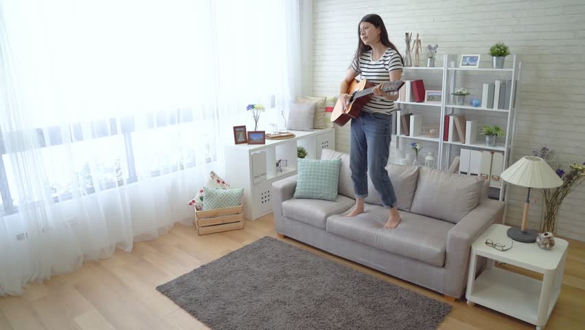 Asian woman holding her wooden guitar and jumped down from the sofa in the living room. She pretends she is a rock star. Royalty-Free Stock Footage #1015315690