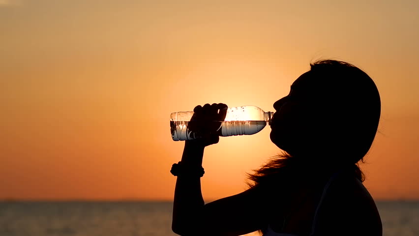 Silhouette of young asian woman drinking water bottle with sunset sky and sea background. Thirsty girl standing on beach is drinking fresh water to replacement on hot days in summer in slow motion. Royalty-Free Stock Footage #1015317664