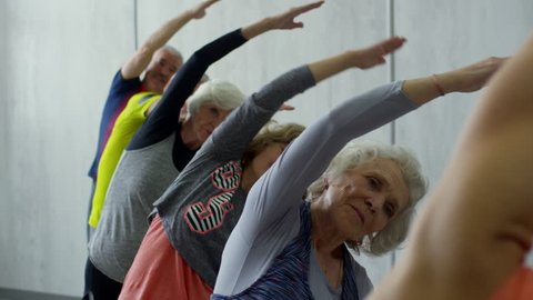 PAN of female yoga instructor and group of senior people standing in line and doing side bends with spread arms during class