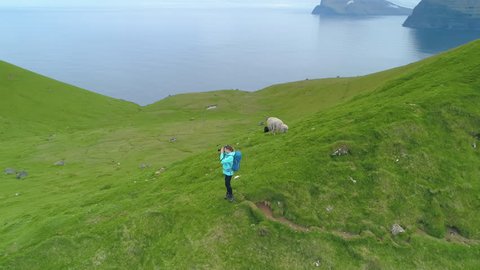 AERIAL: Woman photographer hiking near the Kallur lighthouse taking photos of the breathtaking ocean off a grassy mountain. Flying around young female tourist photographing the scenic countryside.