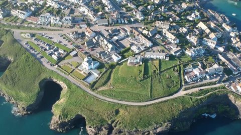 Port Isaac in Cornwall seen by air in upward panning shot revealing the town, blue sea, countryside and coastline behind. Features vehicles and people moving around the town