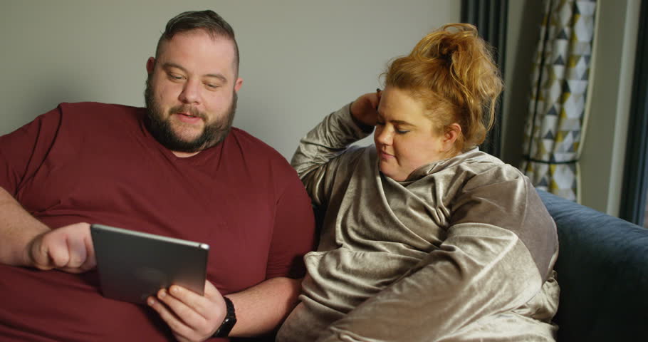 4K Happy overweight couple at home, browsing the internet on computer tablet