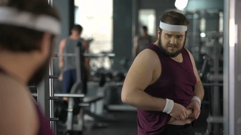 Unhappy overweight man looking at his mirror reflection in gym, diet and sport
