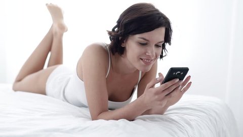 Woman With Mobile Phone Lying On Bed With White Linens