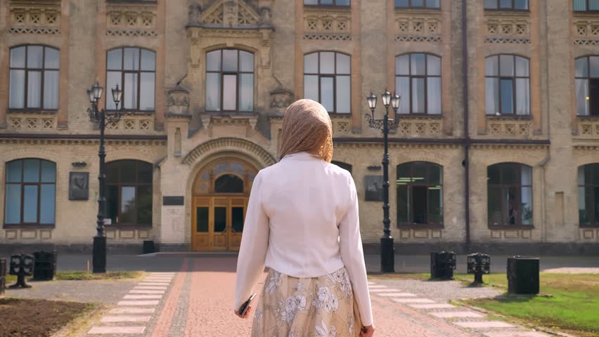 Back view of young muslim woman in hijab walking towards building, looking around, sunny day outdoors