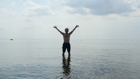 Joyful man standing topless in sea water and waving hands greetings, 4k shot