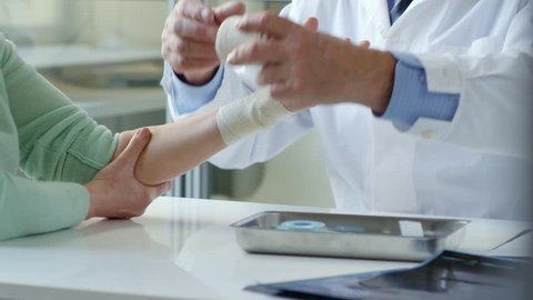 Tilt up of senior male doctor unwrapping elastic bandage from wrist of female patient and telling her to move fingers after recovery