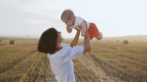 Beautiful woman in a light dress raises high up her charming baby boy and starts to around and smiles. Happy young woman spending time playing with son in the field at sunset, slow motion