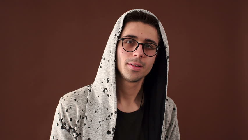 A fashionable young guy in a hood and glasses points a finger at you and winks one eye in the studio on a brown background. Slow motion.
