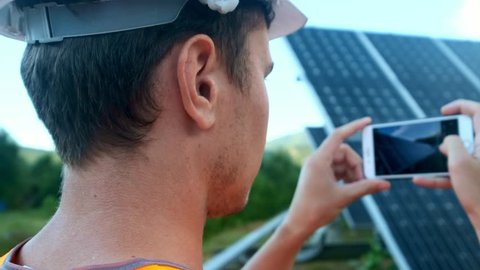 Engineer expert in solar energy photovoltaic panels with remote control performs routine actions for system monitoring using clean, renewable energy. Makes photos of solar panels on a smartphone