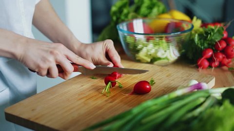 Woman hands with knife cutting radishes for salad. Close up fresh vegetables on kitchen table. Housewife cooking natural and healthy meal on wooden board. Seasonal vegetables ingredients for salad