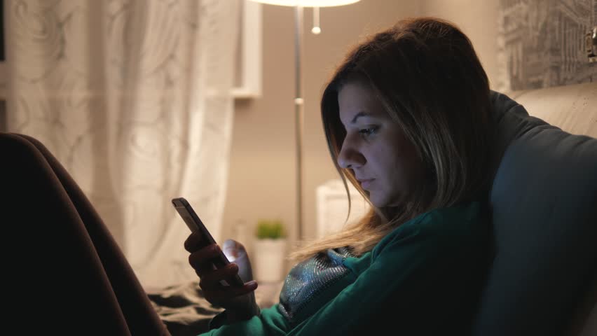 Young beautiful woman lies in bed at home. Uses applications or writes a message in the smartphone. In the evening before going to bed by the light of the lamp, 4k