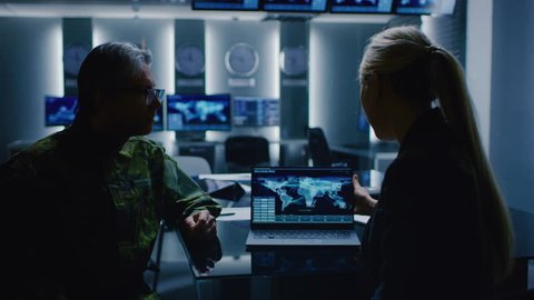 Female Special Agent Talks To Military Man in the Monitoring Room. In the Background Busy System Control Center with Monitors Showing Data Flow. Shot on RED EPIC-W 8K Helium Cinema Camera.