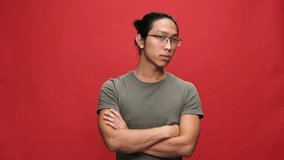 Displeased asian man in t-shirt and eyeglasses with crossed arms waving his head and looking at the camera over red background